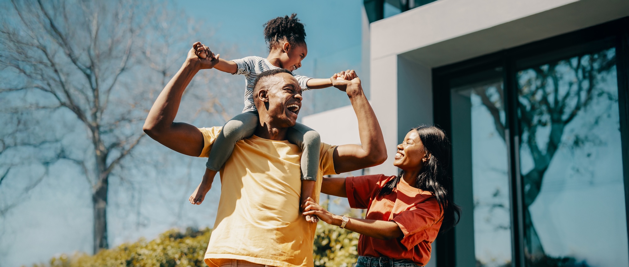 A smiling family outside their home, with a child on her father’s shoulders
