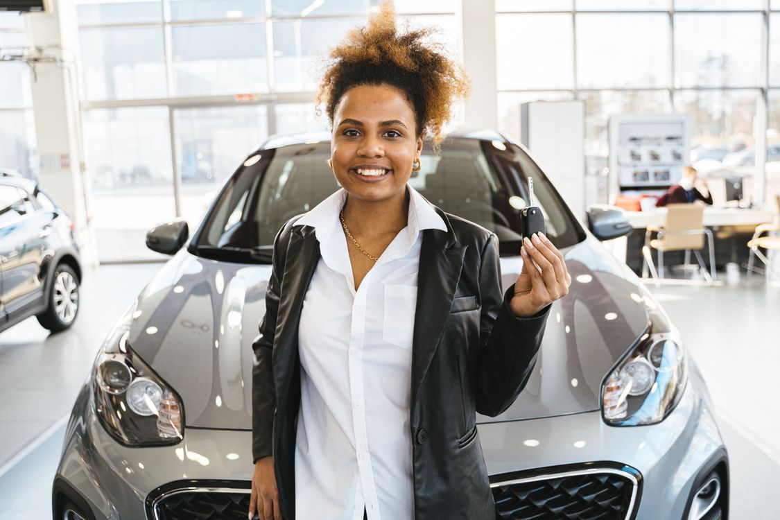 A smiling woman holding car keys beside a car in a showroom.